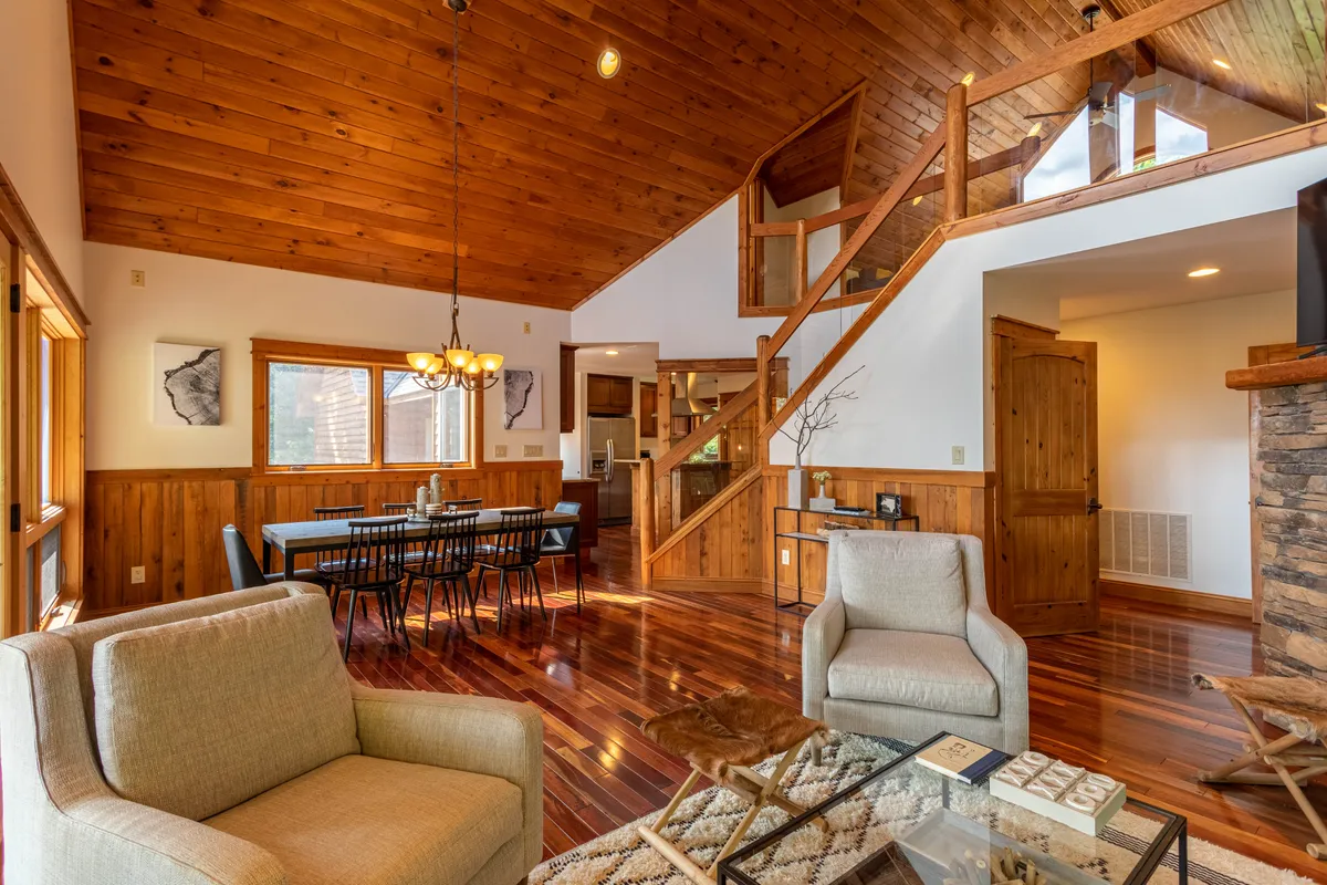 This image captures a wide view of the open-concept main living area, featuring comfortable living room seating, a large wooden dining table, and a glimpse of the wooden ceiling. Expansive windows bring in natural light and offer mountain vistas, while the space feels connected and welcoming, perfect for group gatherings.