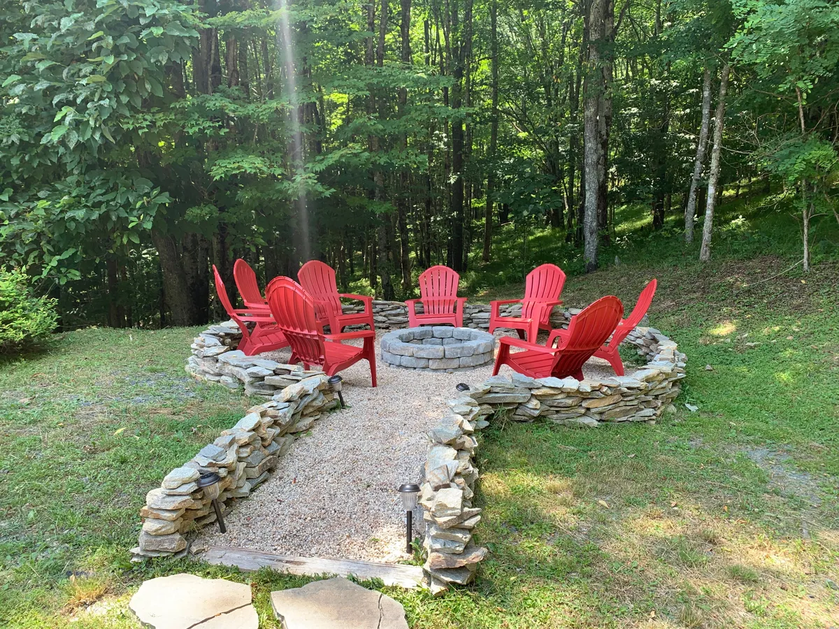This image focuses on a cozy stone fire pit surrounded by red Adirondack chairs, nestled among trees with a gravel path leading to it. The setting is perfect for evening gatherings, storytelling, and stargazing, creating a sense of warmth and togetherness.