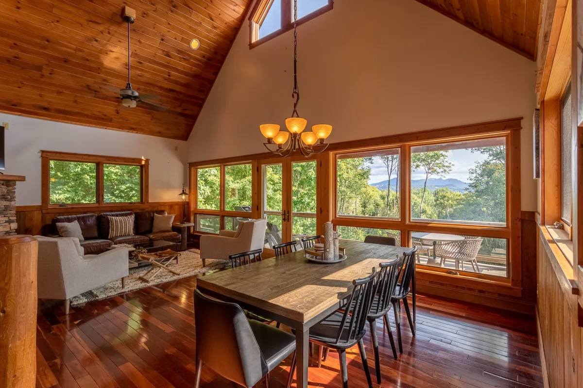 The image shows a bright, airy interior with towering windows framing sunrise views of the surrounding trees. A spacious dining table invites gatherings, surrounded by plush seating and rustic wood beams that accentuate the cabin’s modern mountain charm.