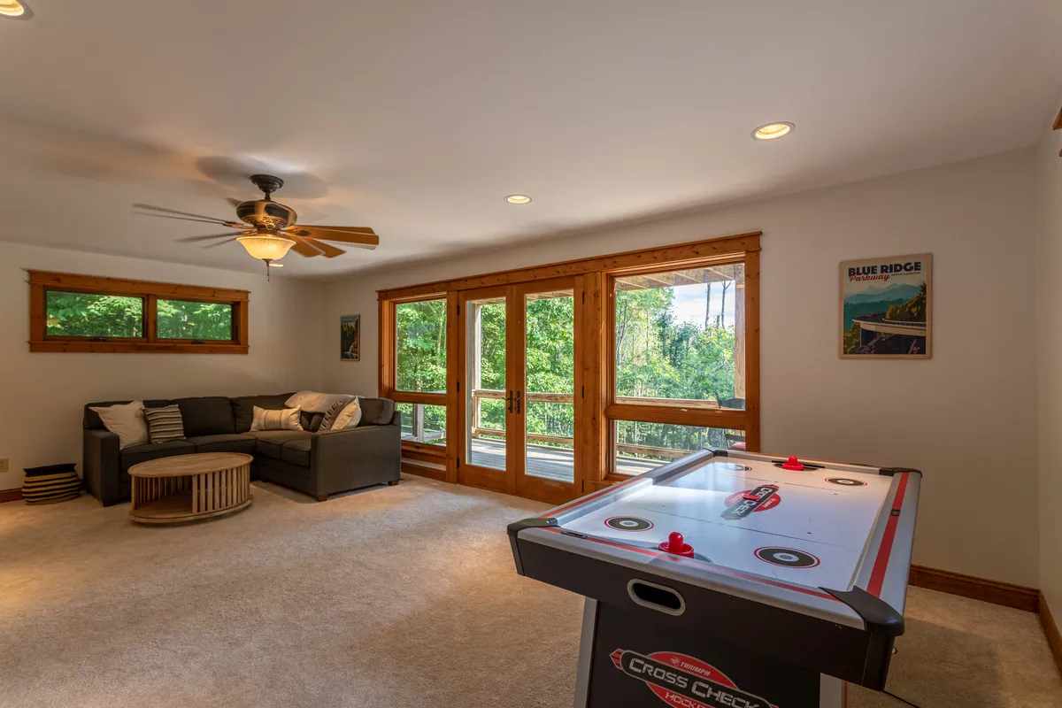 A spacious interior featuring a large sectional sofa, an air hockey table nearby, expansive windows inviting natural light, and a ceiling fan overhead. The room exudes a playful yet cozy vibe, ideal for both games and relaxation.