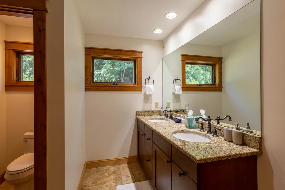 A modern bathroom with double sinks atop a granite countertop, framed by natural wood and flooded with natural light. The space feels serene and offers ample room for morning routines or evening relaxation.