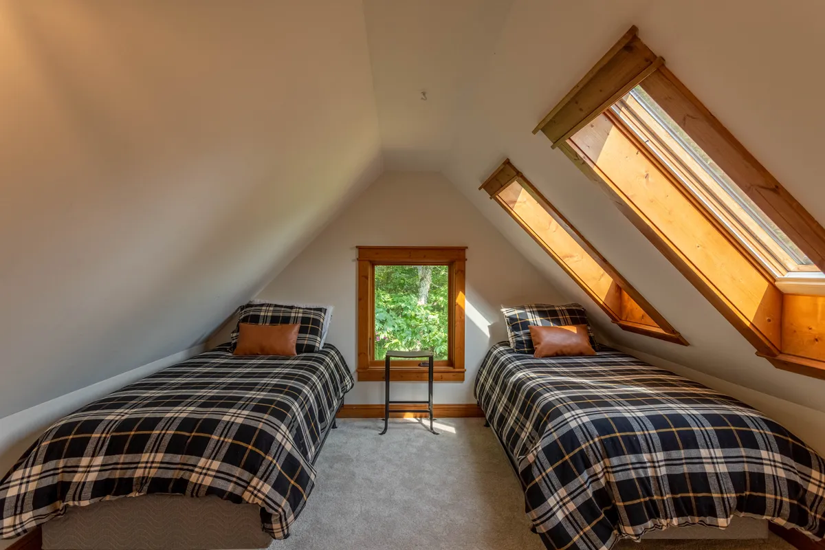 A bright lofted bedroom with two twin beds, warm wooden accents, and large windows that fill the space with morning light. The setting is playful and comfortable, perfect for younger guests or friends seeking a cozy hideaway.