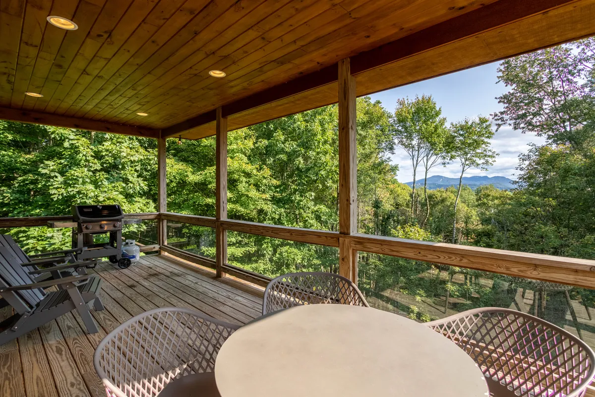 The image showcases a spacious wooden deck area, featuring a grill, table with chairs, and panoramic views of the mountains. The setup suggests opportunities for barbecuing, relaxing, and enjoying nature from the comfort of the deck.