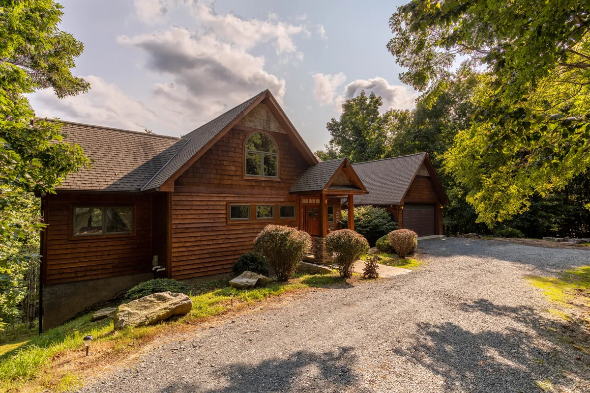 A warmly-lit exterior shot of a wooden home with large windows, surrounded by a landscaped front yard and trees. The porch beckons with comfortable seating, perfect for watching the sunset. The overall mood is serene and inviting, highlighting opportunities for relaxation and outdoor enjoyment.