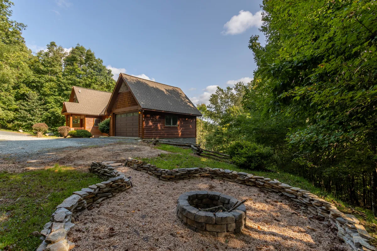 A scenic outdoor area featuring a rustic wooden house framed by trees, with a stone-lined fire pit area set up for comfortable social gatherings. The gravel driveway and surrounding woodlands create a tranquil, private atmosphere ideal for evening relaxation or storytelling around the fire.