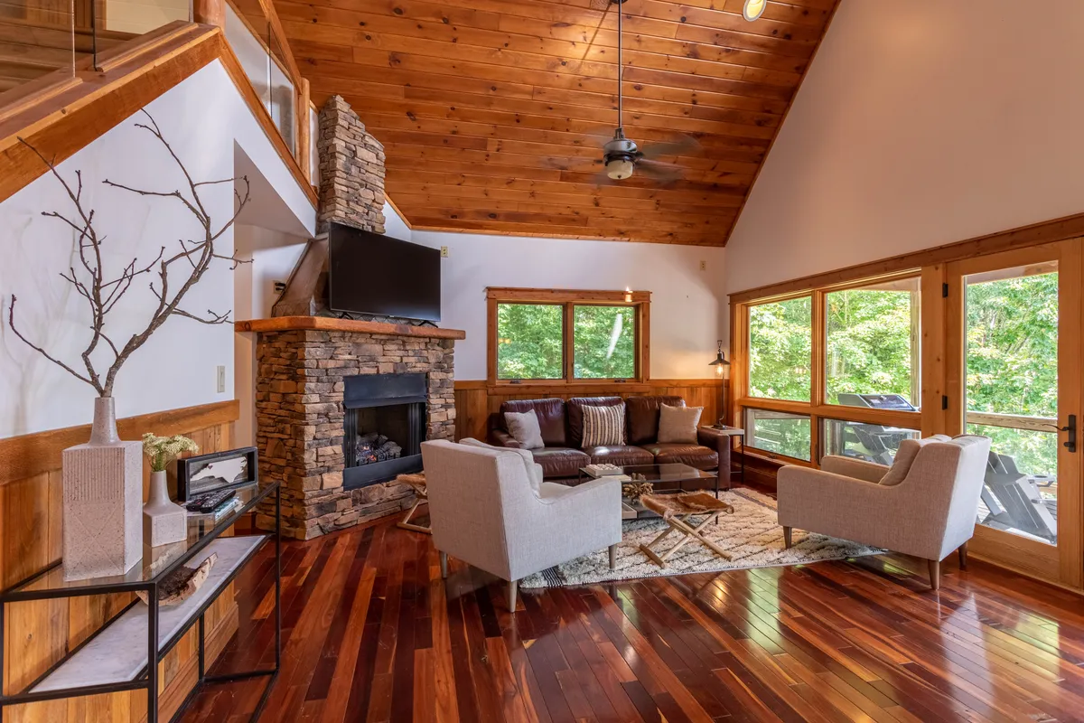 A warm living room scene with a focus on a stone gas fireplace, plush leather seating, and large windows looking out on nature. The space glows invitingly, promising comfort, relaxation, and memorable evenings by the fire.